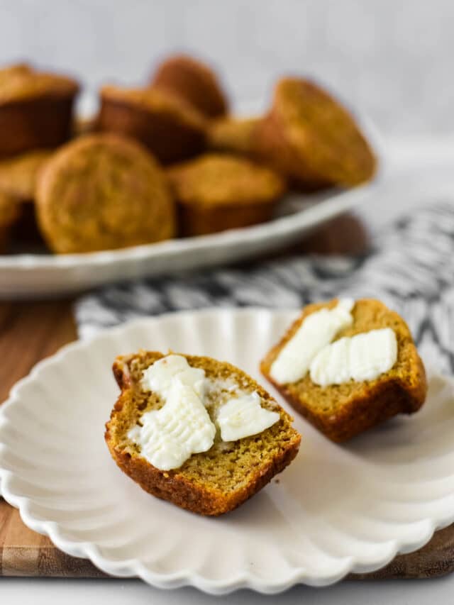 muffin on white plate, in front of platter piled with more muffins. Muffin has been sliced in half and butter smeared on both halves.