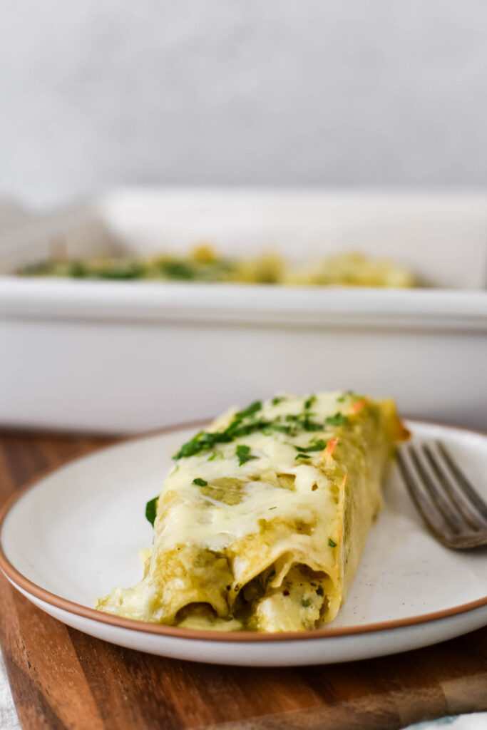 two chicken enchiladas with green chili and sour cream on white plate with fork side and baking dish in background