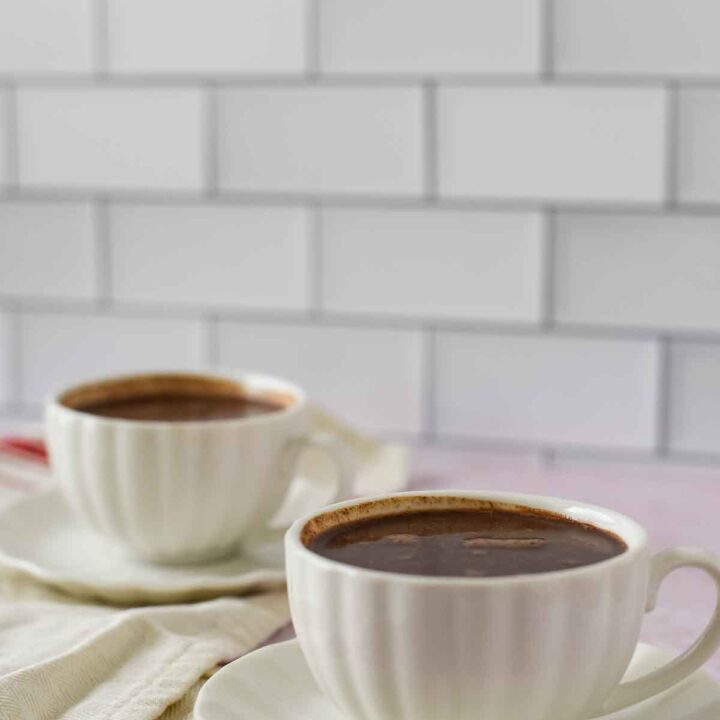 two white mugs of hot chocolate sitting on white saucers. Mugs are on pink marble with white subway tile behind