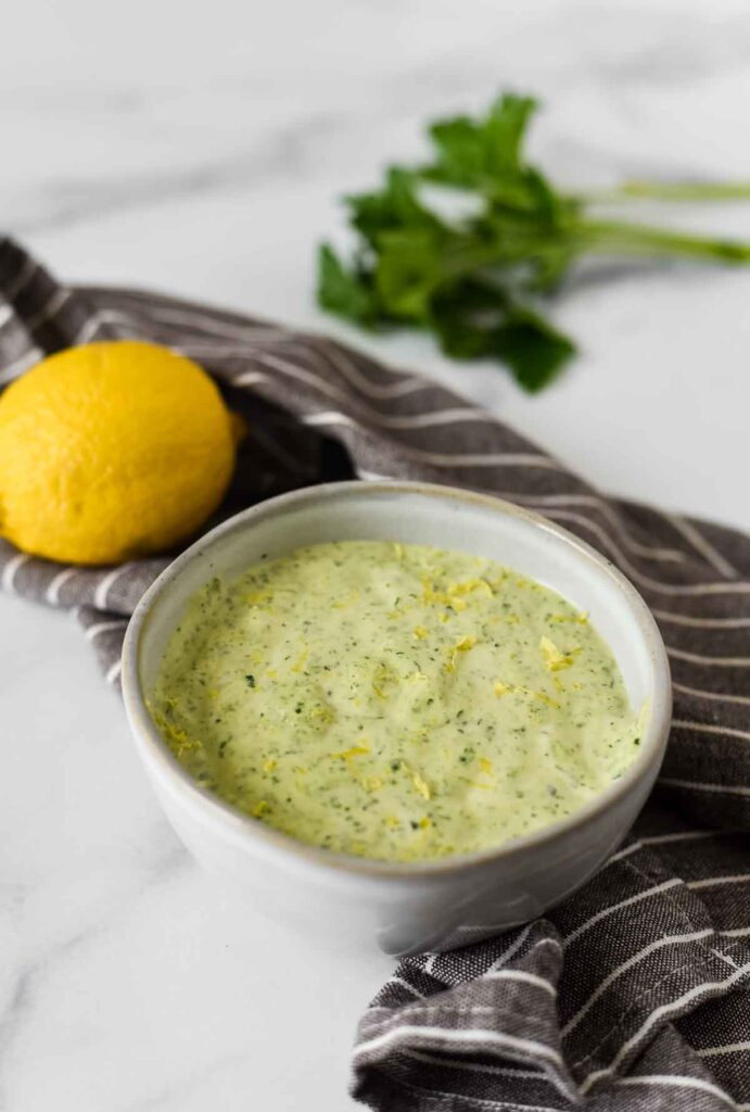 lemon herb tahini sauce in bowl wtih lemon zest on top, with parsley and whole lemon in background