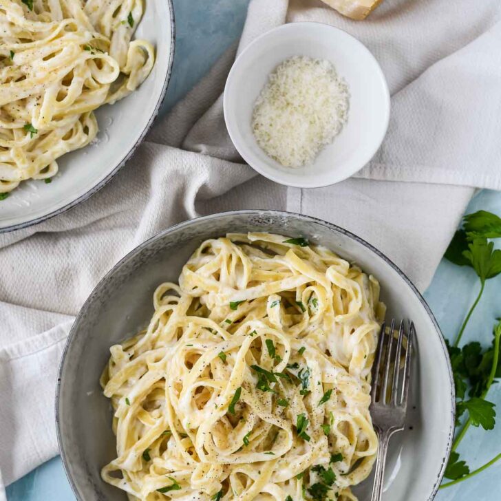 two plates with pasta and Alfredo sauce, next to cheese and napkin