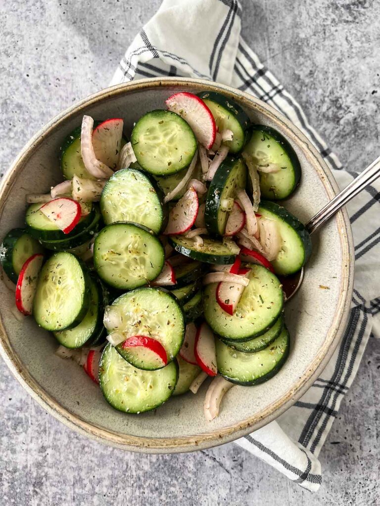 grey serving bowl with sliced cucumbers, radishes and shallots with vinaigrette dressing