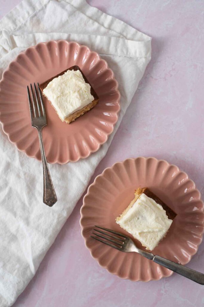 overhead view of two pieces of vanilla sourdough cake with vanilla frosting on pink plates with forks, and white napkin diagnoally on counter