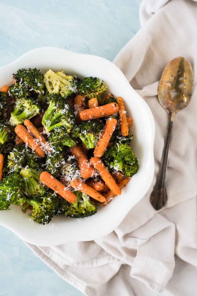 overhead view of white serving bowl filled with roasted broccoli and carrots and silver spoon beside