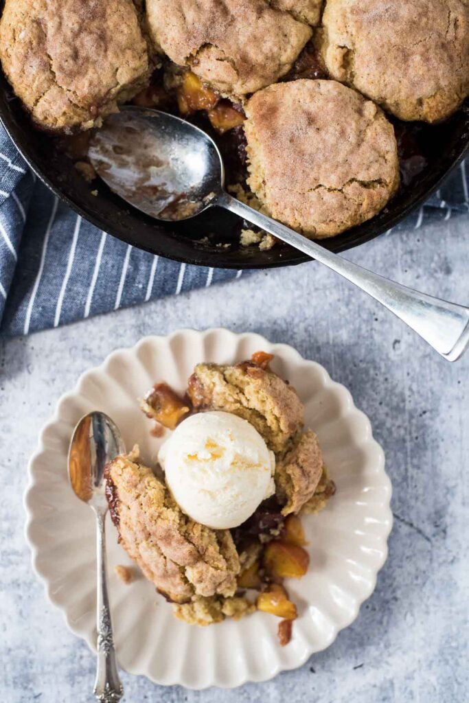 white plate wtih serving of peach cobbler and vanilla ice cream next to cast iron skillet with more cobbler and serving spoon