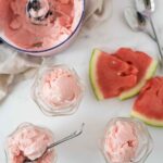three glass bowls of watermelon whip with spoons and slices of watermelon thrown around, next to a food processor bowl with more whip