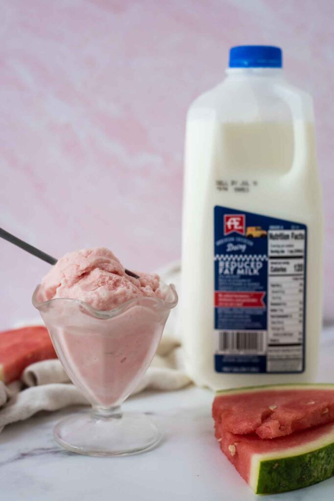 glass of watermelon whip with spoon next to slice of watermelon and half gallon of AE dairy milk
