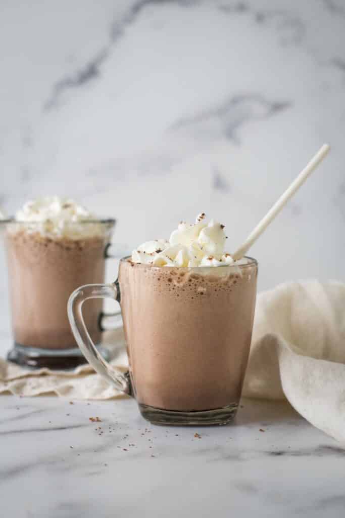 two glass mugs with hot chocolate, whipped cream, and white paper straws