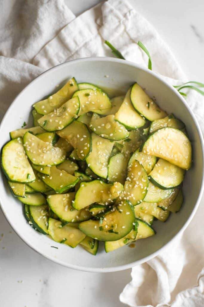 white bowl with sautéed zucchini cut into half moon shapes, sprinkled with sesame seeds and green onions