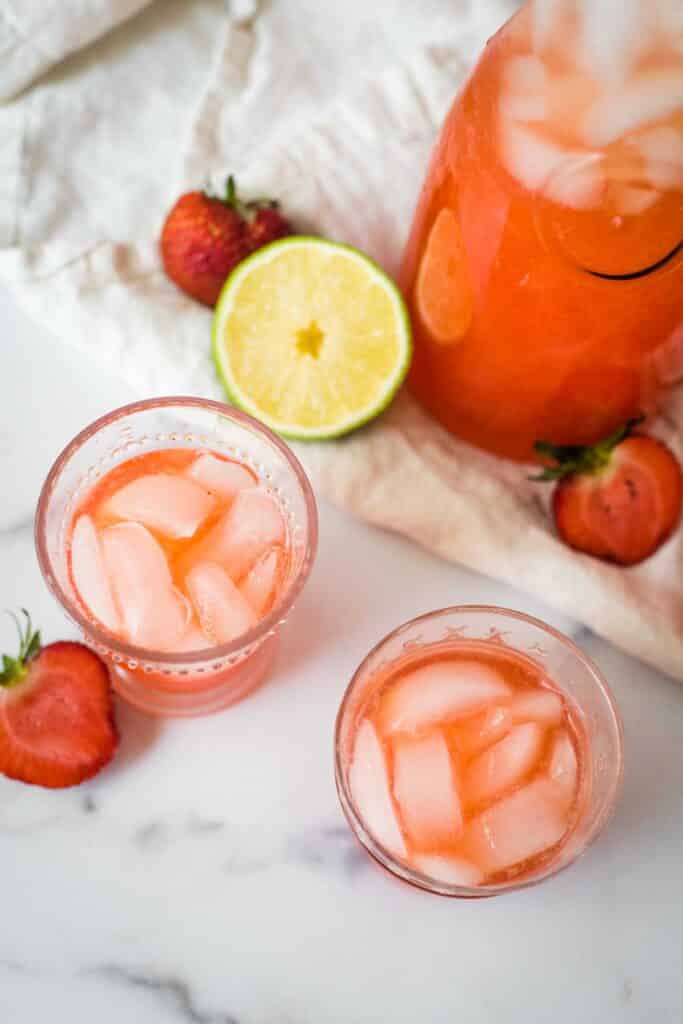 overhead view of two glasses filled with strawberry limeade and ice beside pitcher and sliced limes