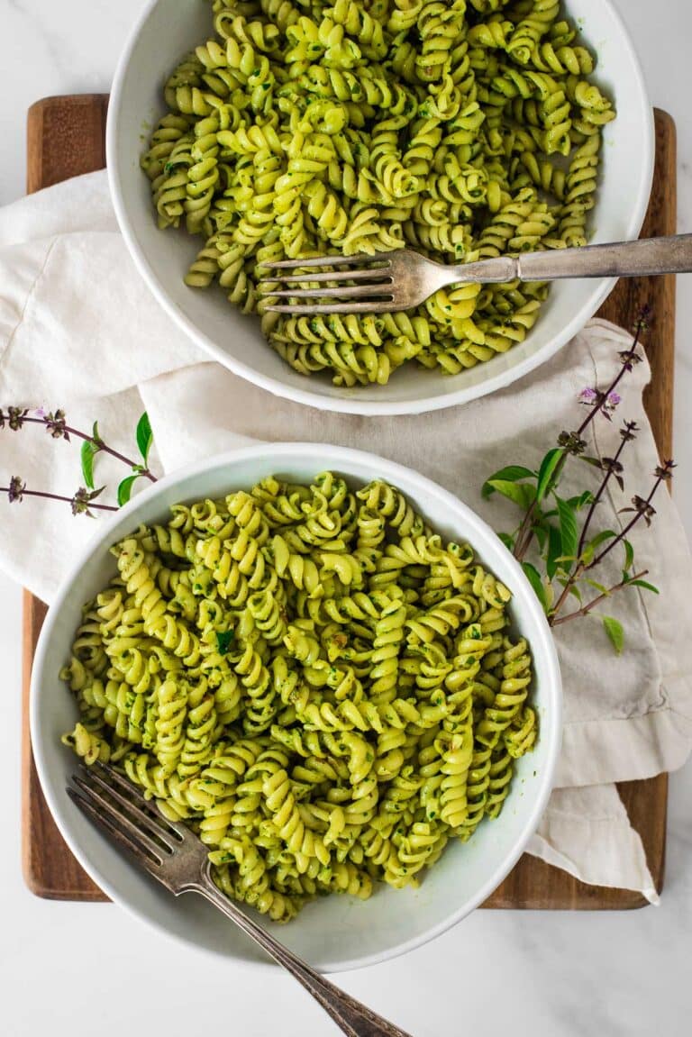 two white bowls of pasta with thai basil pesto and forks, thai basil flowers around bowls