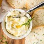 overhead view of small bowl with whipped butter topped with honey and fresh rosemary, with small knife in bowl and surrounded by bread slices