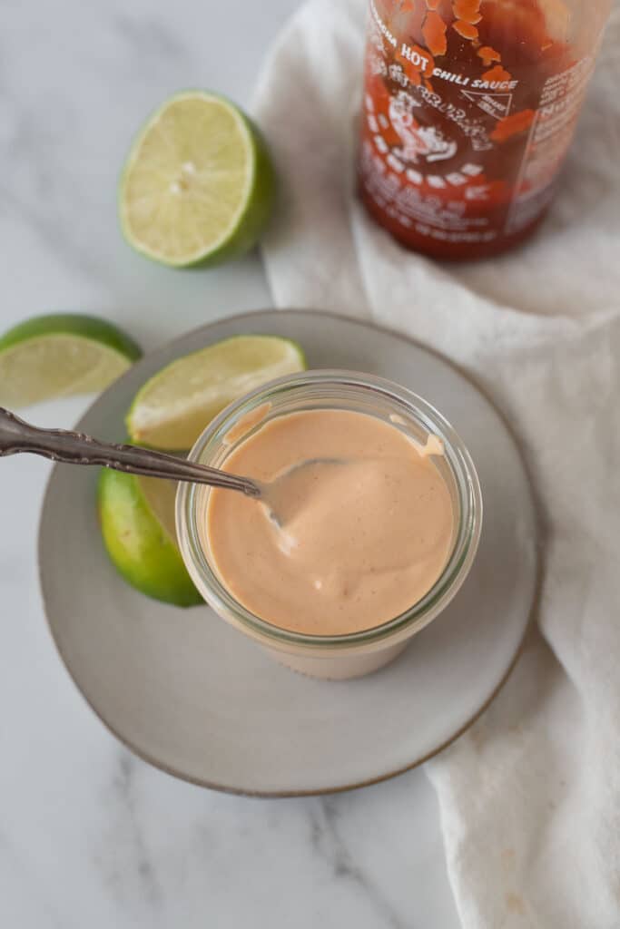 overhead view of glass container of spicy mayo and spoon, with limes and sriracha in background