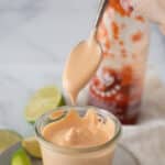 glass container filled with spicy mayo, with spoon taking some out and limes behind