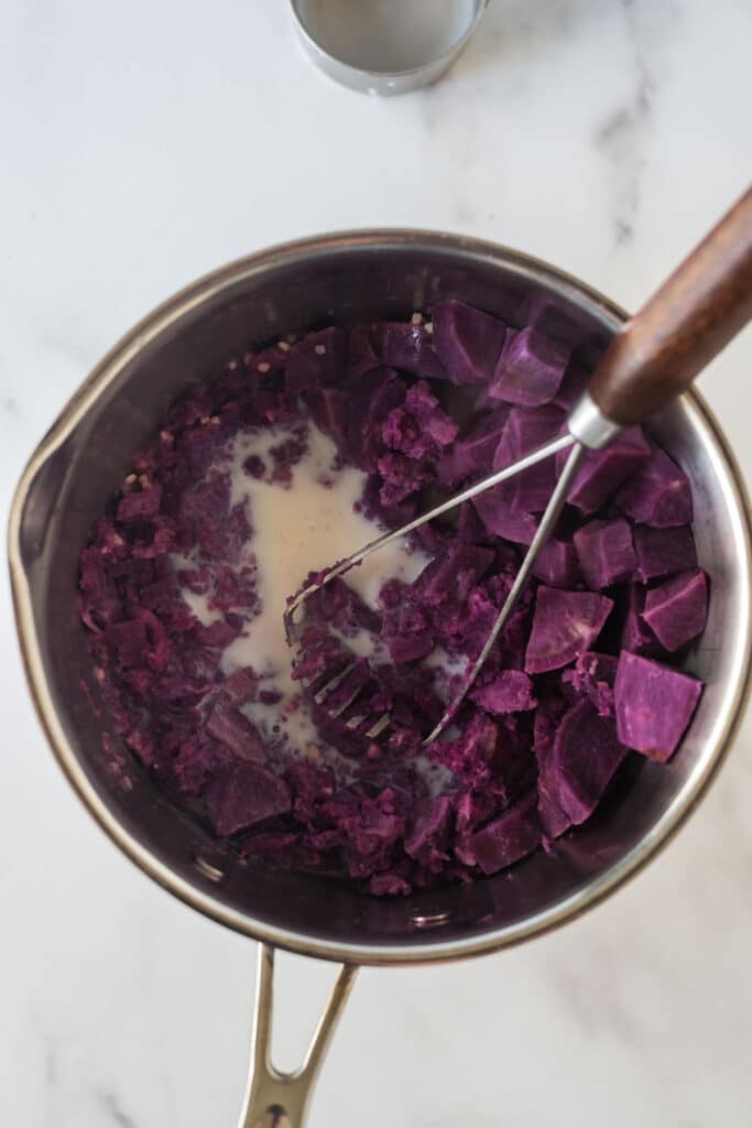 saucepan with cooked purple potatoes cubes, milk, and garlic with potato masher