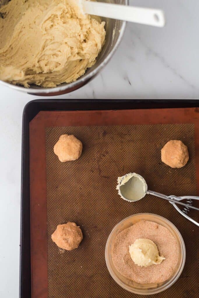 cookie dough being scooped from bowl and rolled in cinnamon sugar  with baking pan