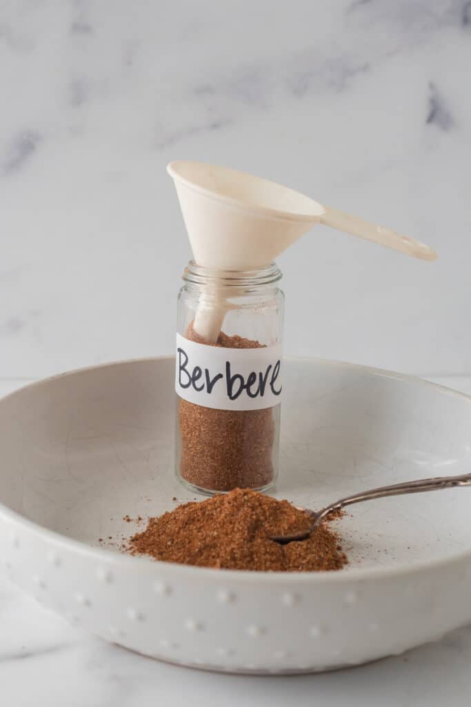 a glass jar of berbere spice mix in a shallow white bowl. A funnel is inside the jar and a spoon is in the spice mix in the bowl.