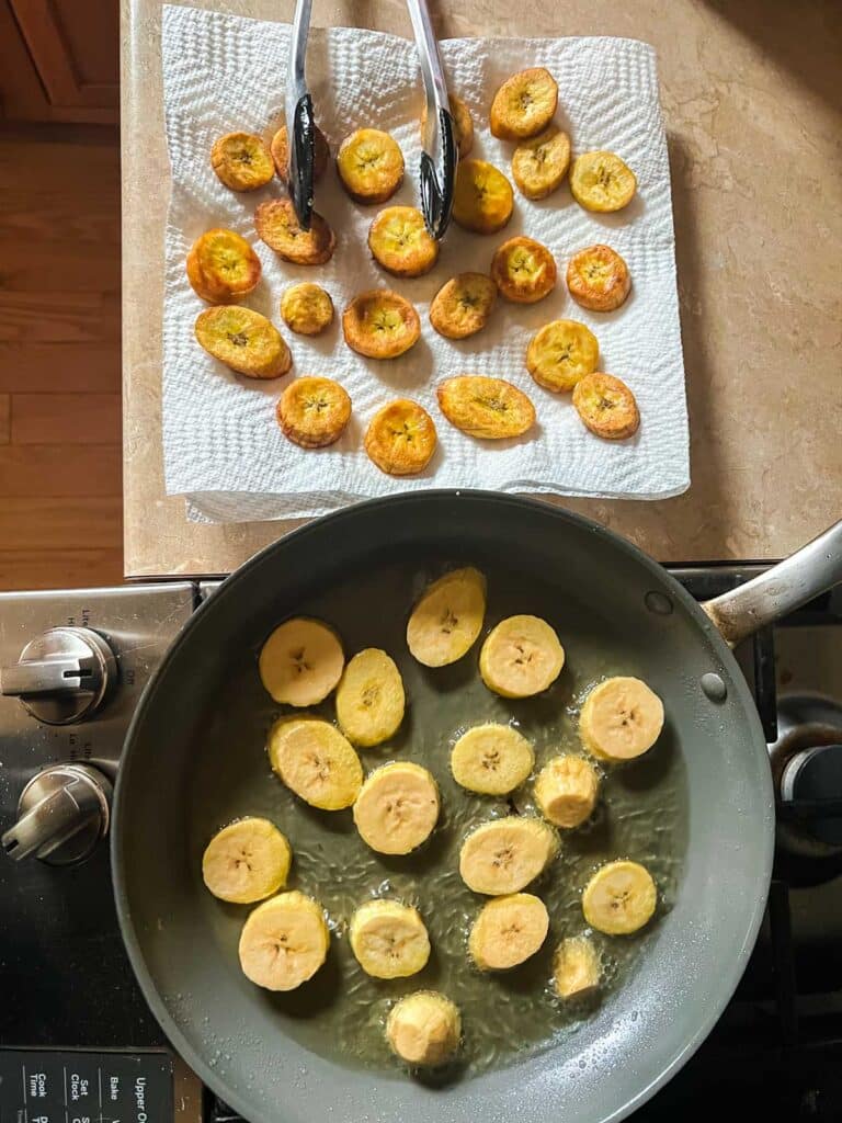 skillet where plantain slices are frying next to pepper towel lined plate where freshly fried plantains are resting