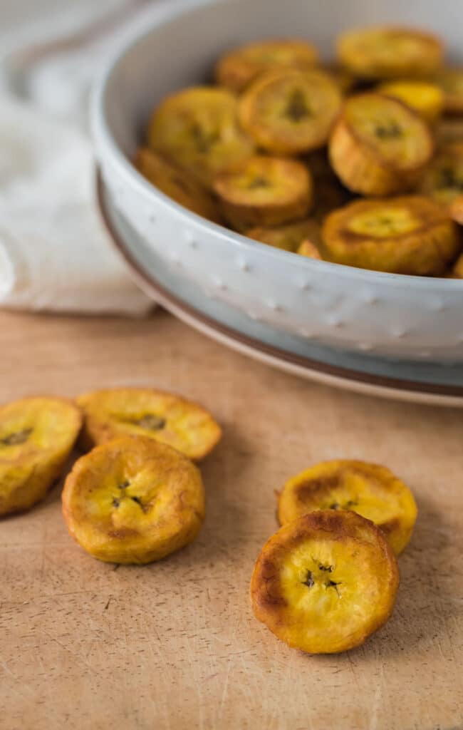 fried plantain slices on wooden board with bowl of more behind