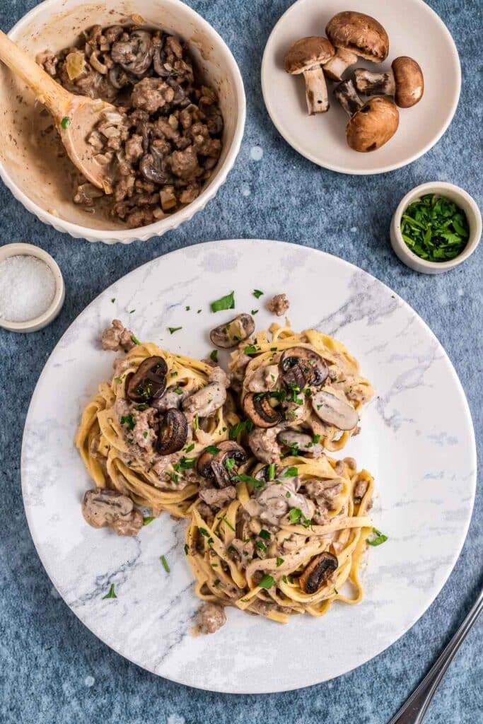 overhead view of white plate with beef stroganoff with bowls of mushrooms beside