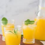 three glasses of mango lemonade on wooden serving board with pitcher behind