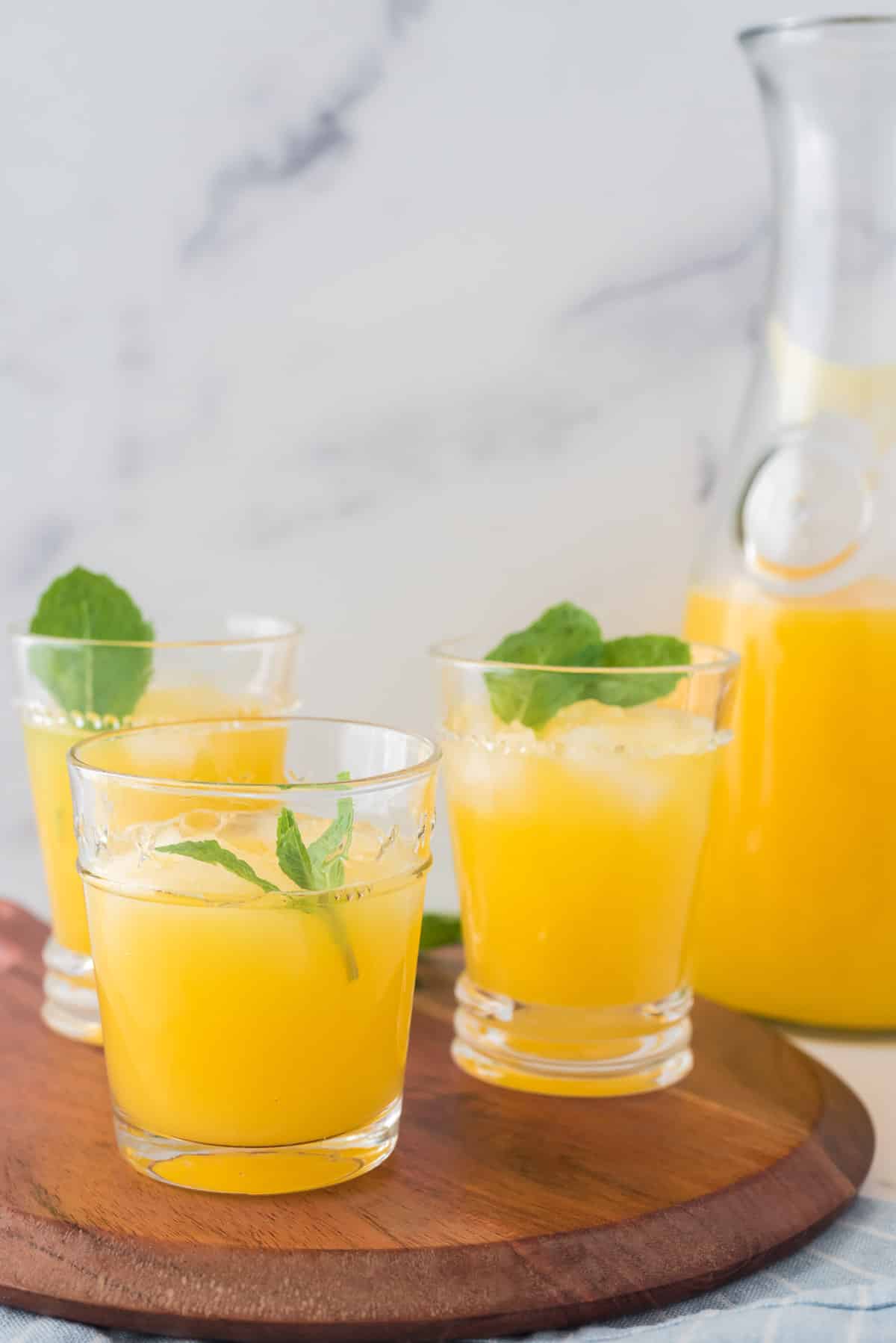 three glasses of mango lemonade on wooden serving board with pitcher behind