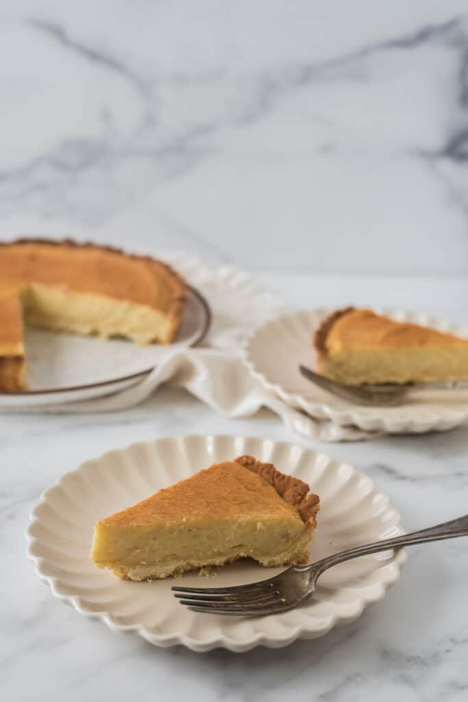 two slices of milk tart on white plates with forks, and rest of tart in background