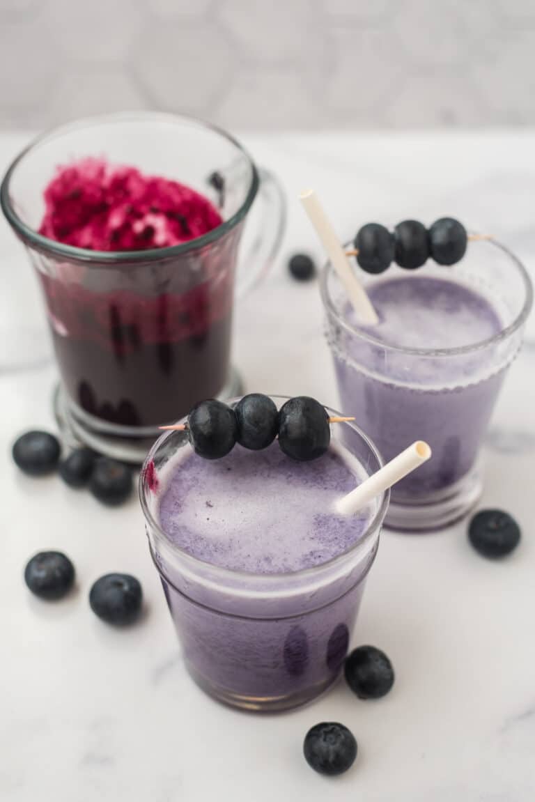 two glasses of blueberry milk with white straws next to glass of blueberry syrup, with fresh blueberries scattered around all three glasses