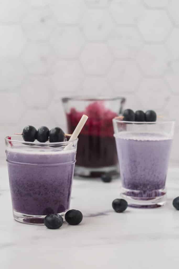two glasses of blueberry milk with white straws and pitcher of blueberry syrup behind 
