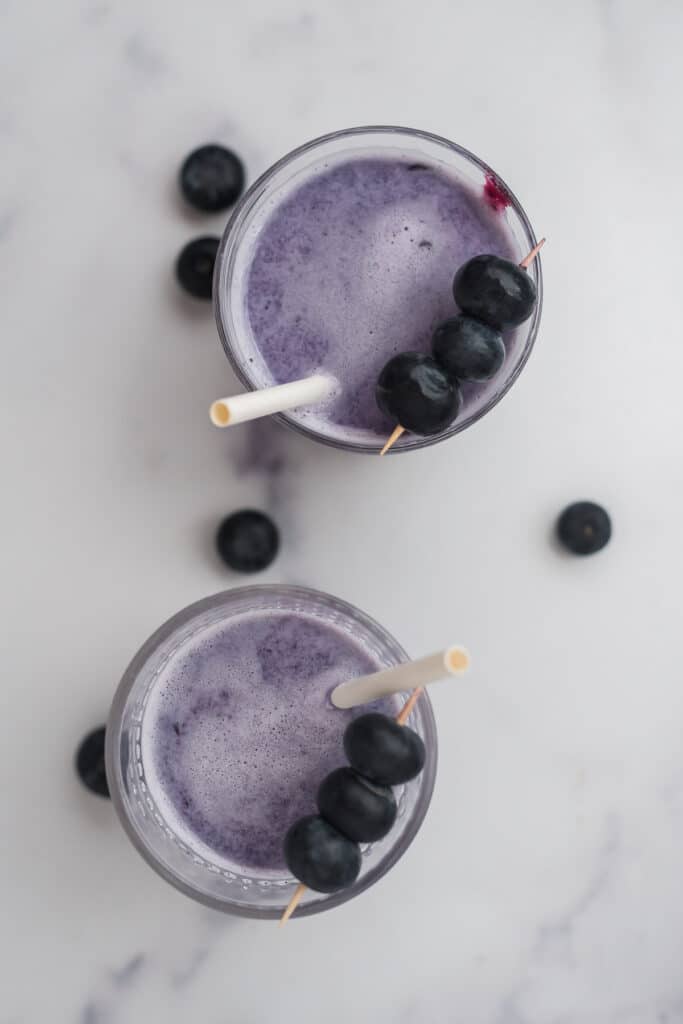 overhead image of two glasses filled with blueberry milk, with white straws and garnishes of fresh berries