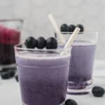 two glasses with purple blueberry syrup and milk mixed together, with white straws and fresh blueberry skewers are garnishes. Fresh blueberries scattered around counter and glass pitcher of blueberry syrup in background