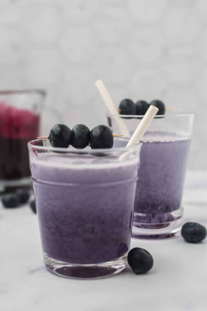 two glasses with purple blueberry syrup and milk mixed together, with white straws and fresh blueberry skewers are garnishes. Fresh blueberries scattered around counter and glass pitcher of blueberry syrup in background