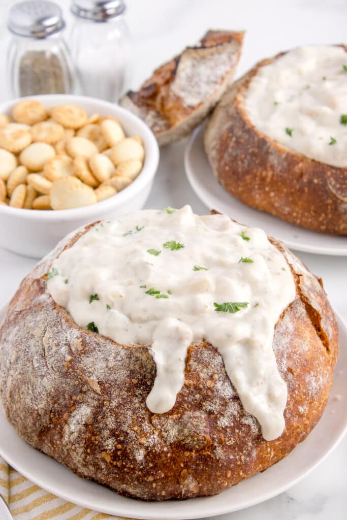 two sourdough bread bowls on plates, filled with chowder and soup crackers behind