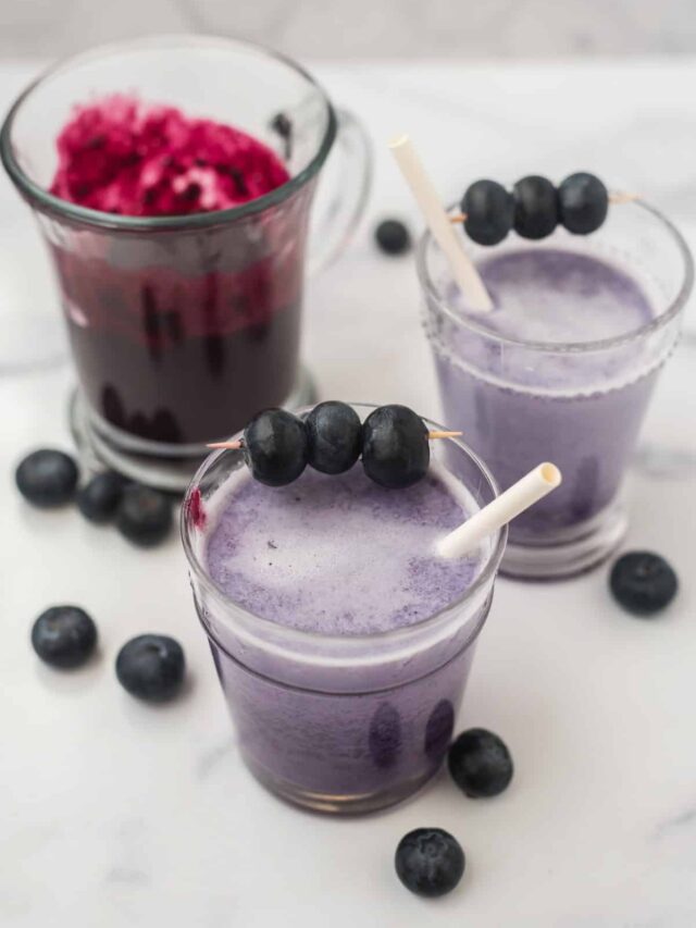 two glasses of blueberry milk with white straws next to glass of blueberry syrup, with fresh blueberries scattered around all three glasses