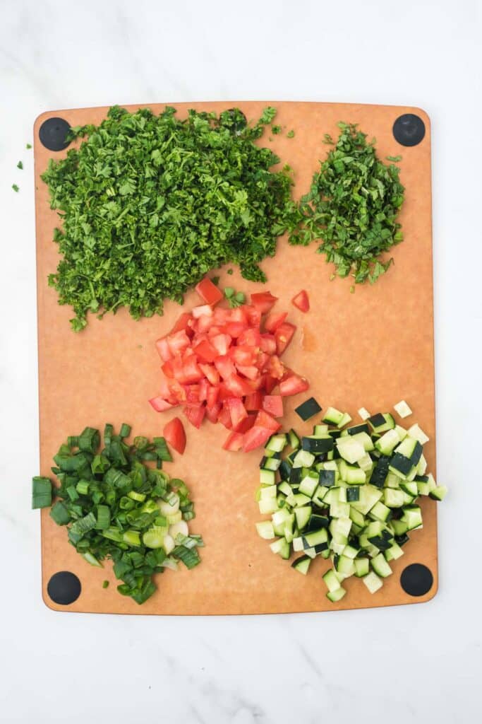 cutting board with chopped parsley, chopped mint, chopped tomato, chopped green onion and chopped cucumber