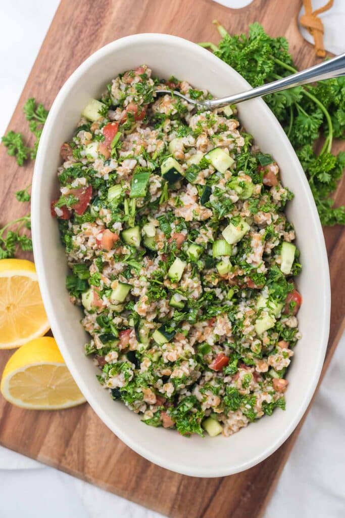 overhead view of tabbouleh in white serving bowl with spoon, cut lemon and fresh parsley tucked around bowl
