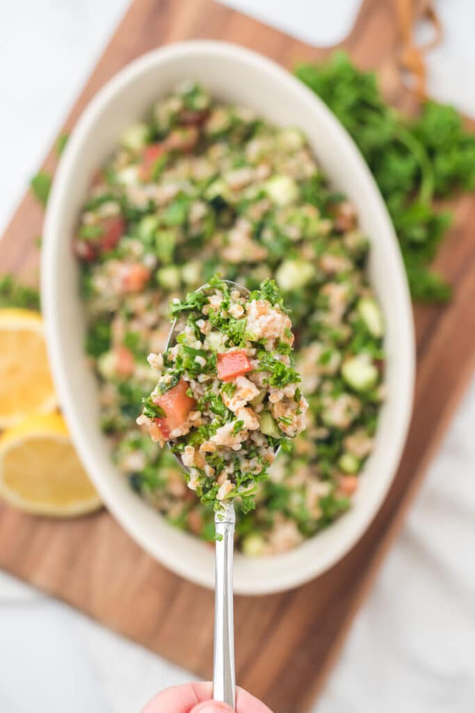 overhead view of tabbouleh in white serving bowl on cutting board, with filled serving spoon up above and in focus