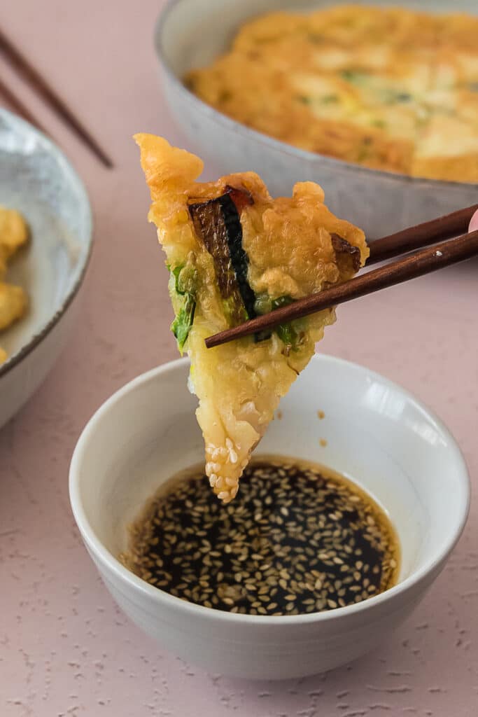 chopsticks holding piece of korean pancake into bowl of dipping sauce