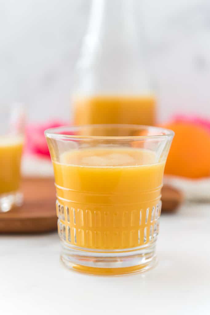 small glass filled with pot juice, with a pitcher and orange blurred in background