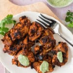 overhead view of white platter on cutting board with grilled chicken, some of the pieces dotted with green sauce, with serving fork on side of platter and bowl of green sauce in corner of photo