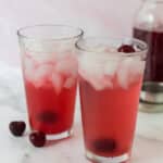 two glasses with cherryade and ice with cherries surrounding them and jar of cherry syrup in background