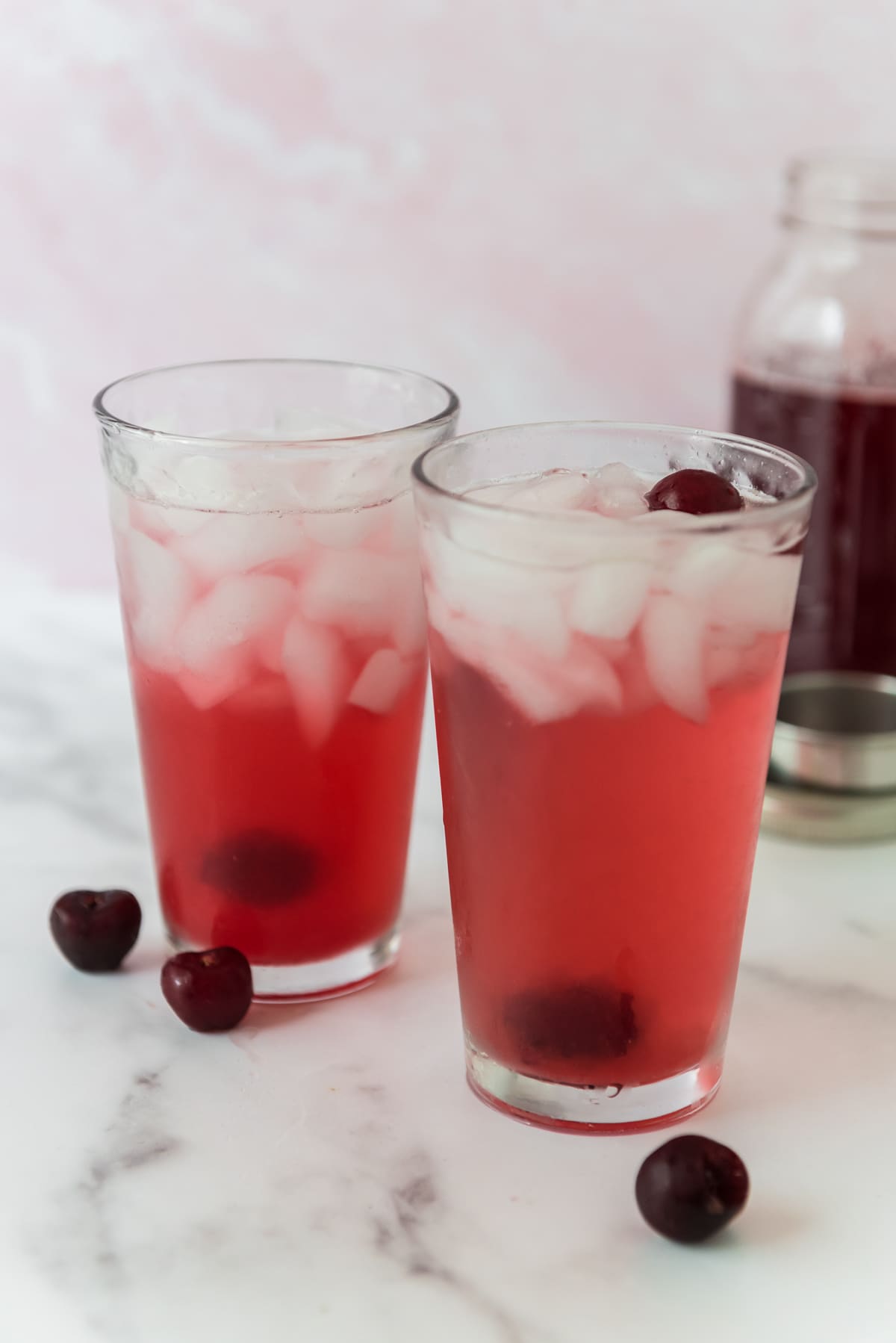 two glasses with cherryade and ice with cherries surrounding them and jar of cherry syrup in background