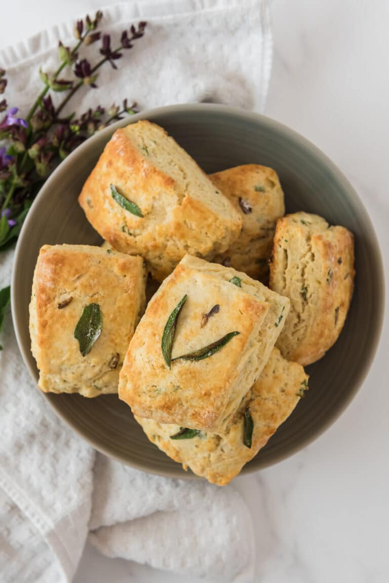 overhead view of grey bowl filled with biscuits topped with sage leaves, and a bundle of fresh sage with purple flowers beside bowl