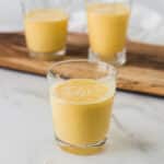 three small glasses with mango milkshake, one in front and in focus and two in background on wooden serving tray