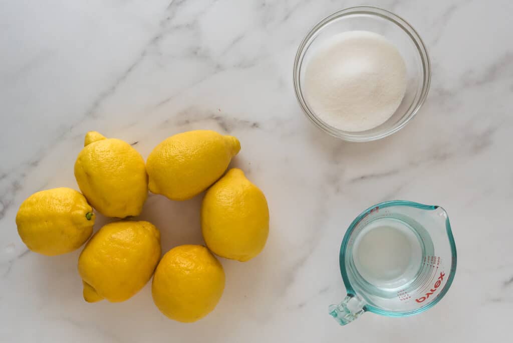 overhead view of marble counter with 6 lemons, glass bowl of sugar and glass measuring cup of water