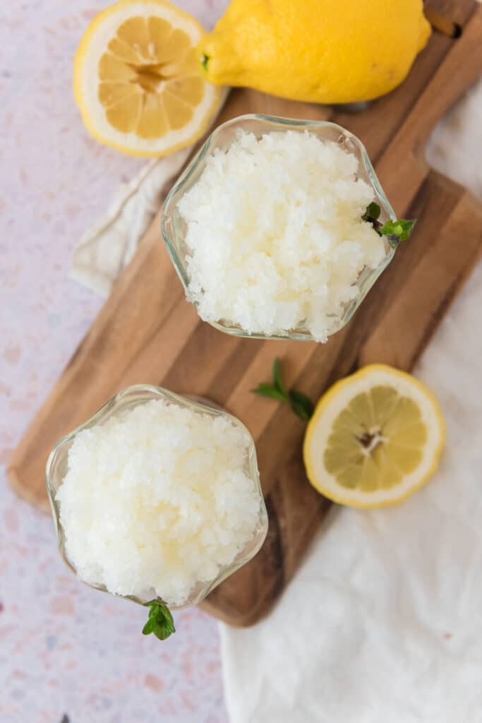 overhead view of two glass bowls with lemon granita garnished with sprigs of mint, and surrounded by lemon slices