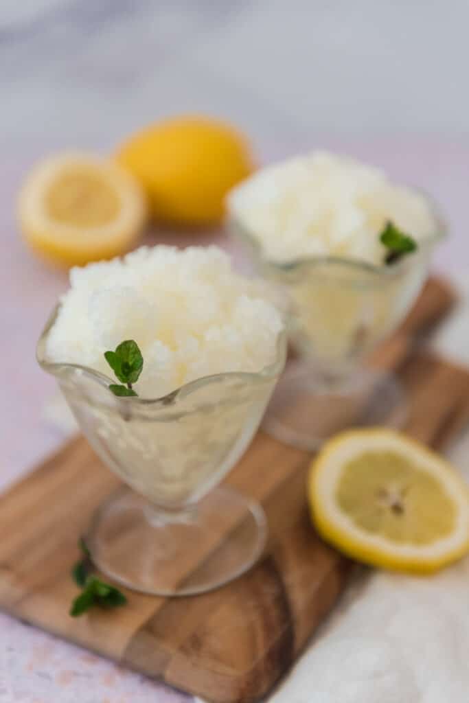 two glass bowls with stems filled with lemon granita and garnished with small sprigs of mint, with lemon slices around
