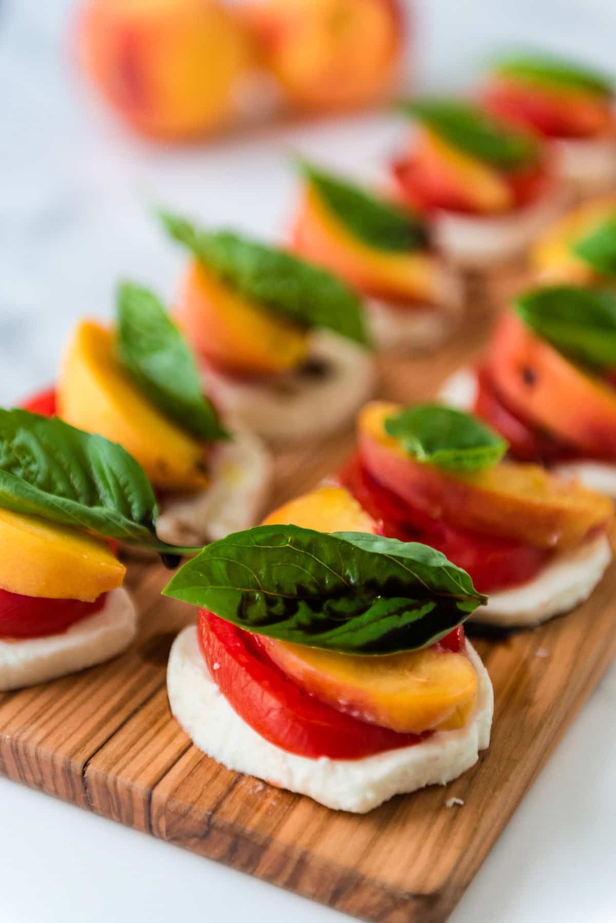 closeup view of wooden cutting board with stacks of mozzarella, tomatoes, peaches, and basil drizzled with balsamic vinegar