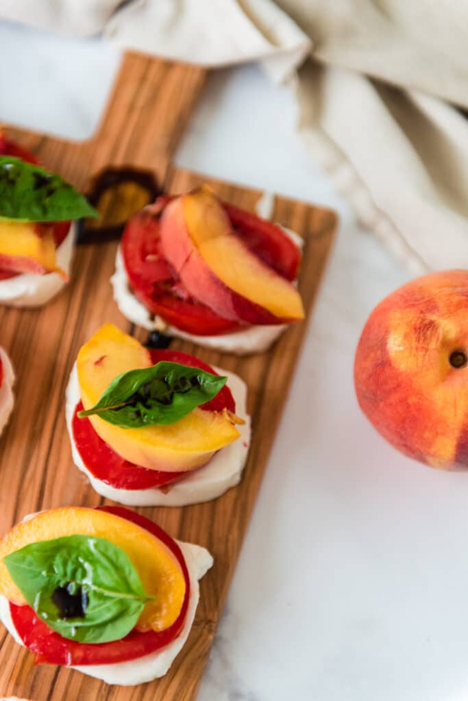 stacks of mozzarella, tomatoes, peaches and fresh basil on wooden cutting board