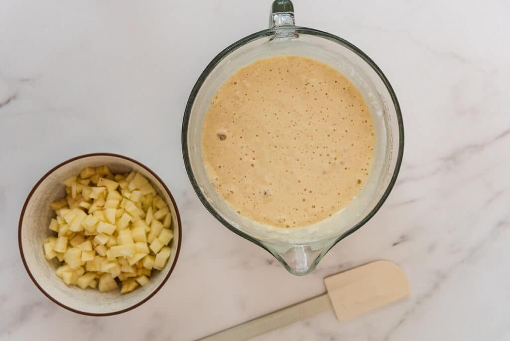 overhead view of yeasted pancake batter in glass mixing bowl with diced apples in ceramic bowl